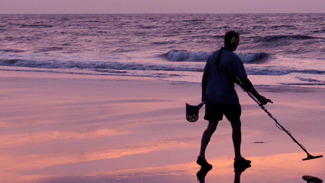 Person som går med metalldetektor på stranda i solnedgang
