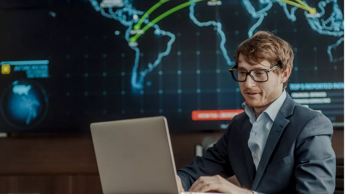 Mann sitter og jobber på PC med verdenskart på skjerm i bakgrunnen